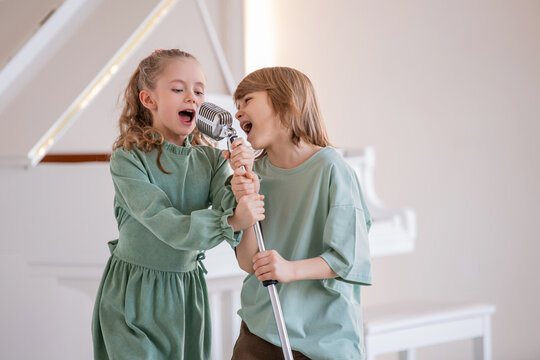 Two children sing into a microphone against the backdrop of a white piano.