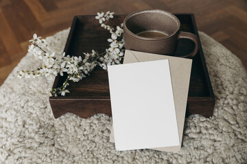 Spring Easter still life. Greeting card, envelope mockup. Cup of coffee. Feminine photo. Floral scene. Blurred cherry tree blossoms on wool taburet, stool. Defocused wooden parquet floor. Front view.