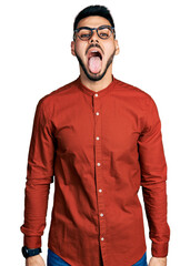 Young hispanic man with beard wearing business shirt and glasses sticking tongue out happy with funny expression. emotion concept.