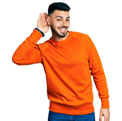 Young hispanic man with beard wearing casual orange sweater smiling with hand over ear listening an...