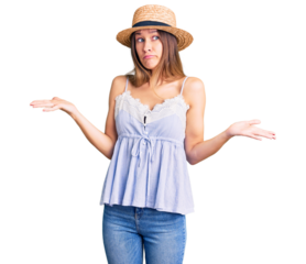 Beautiful brunette young woman wearing summer hat clueless and confused expression with arms and hands raised. doubt concept.