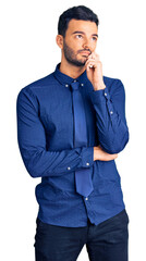 Fototapeta premium Young handsome hispanic man wearing business clothes serious face thinking about question with hand on chin, thoughtful about confusing idea