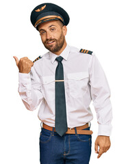 Handsome man with beard wearing airplane pilot uniform smiling with happy face looking and pointing to the side with thumb up. © Krakenimages.com