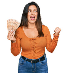 Beautiful hispanic woman holding 50 brazilian real banknotes pointing thumb up to the side smiling...