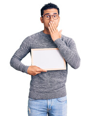 Hispanic handsome young man holding blank empty banner covering mouth with hand, shocked and afraid for mistake. surprised expression