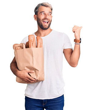 Young handsome blond man holding paper bag with bread pointing thumb up to the side smiling happy with open mouth