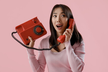 Young surprised Asian woman talking with retro phone on pink background