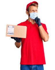 Handsome blond man with beard holding delivery box wearing medical mask serious face thinking about...