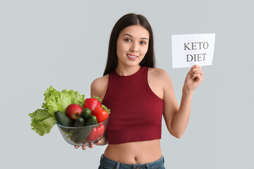Beautiful young happy Asian woman holding paper sheet with text KETO DIET and bowl of fresh vegetables on grey background