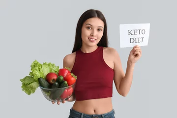  Beautiful young happy Asian woman holding paper sheet with text KETO DIET and bowl of fresh vegetables on grey background © Pixel-Shot
