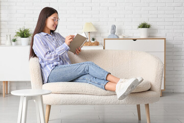 Young happy Asian woman in stylish eyeglasses with book sitting on sofa at home
