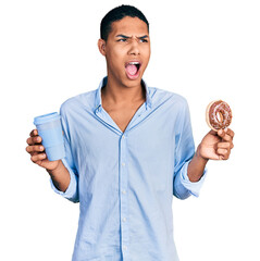 Young hispanic man eating doughnut and drinking coffee angry and mad screaming frustrated and...