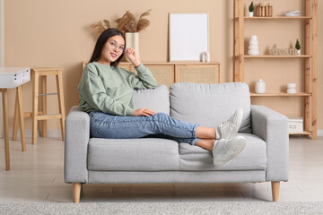 Beautiful young happy Asian woman in stylish eyeglasses sitting on sofa at home