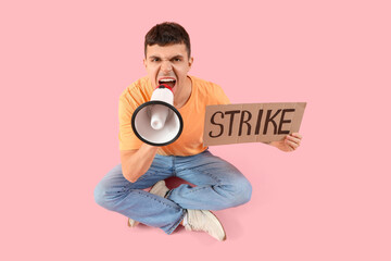 Protesting young man shouting into megaphone and holding placard with word STRIKE on pink background