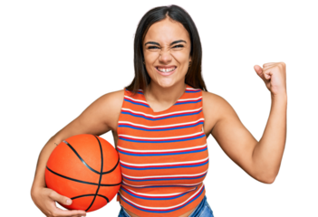 Young brunette woman holding basketball ball screaming proud, celebrating victory and success very excited with raised arms