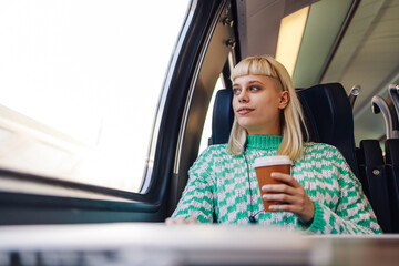 A girl is traveling by train, looking trough window and having coffee.