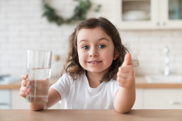 Cute little caucasian girl daughter showing thumb up drinking water, holding a glass of water, hydration, healthy eating habits, daily consumption rate , intake