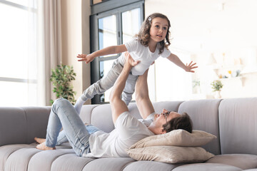 Happy young father and his little daughter playing lying on the sofa, having fun and spending time...