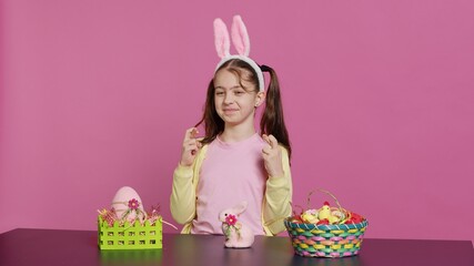 Obraz premium Small cheerful kid posing with fingers crossed in studio, sitting at a table filled with easter festive ornaments. Young schoolgirl hoping for luck and fortune during spring holiday celebration