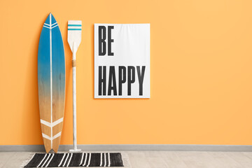 Surfboard, picture and paddle near orange wall in room