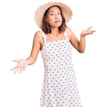 Young beautiful chinese girl wearing summer hat clueless and confused expression with arms and hands raised. doubt concept.