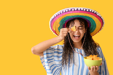 Beautiful young African-American woman in sombrero hat holding bowl with tasty nachos on yellow background