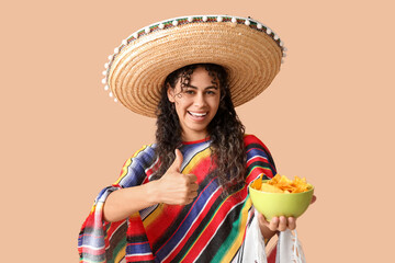 Beautiful young African-American woman in sombrero hat holding bowl with tasty nachos and showing thumb-up gesture on brown background