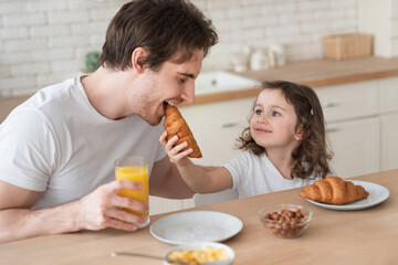 Little cute kid girl daughter feeding his young father with croissant for breakfast spending time...