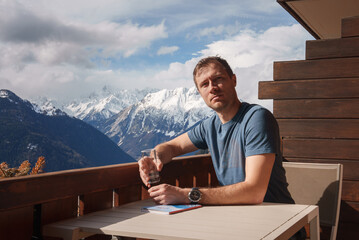 Man sitting at outdoor table on terrace with mountain view holding drink. Wearing blue t shirt,...