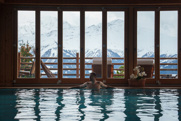 Indoor swimming pool with wooden interior and glass doors showing snow covered mountains. Relaxing setting, potted plants, balcony with outdoor furniture. Serene ambiance. Ideal for relaxation. - obrazy, fototapety, plakaty