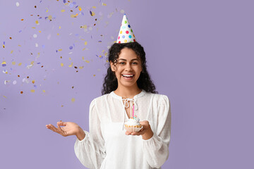 Beautiful young African-American woman in party hat holding birthday cupcake with burning candle...