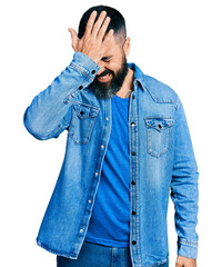 Hispanic man with beard wearing casual denim jacket surprised with hand on head for mistake,...