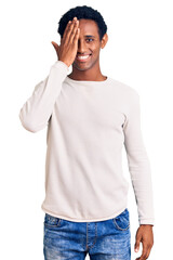 African handsome man wearing casual winter sweater covering one eye with hand, confident smile on face and surprise emotion.