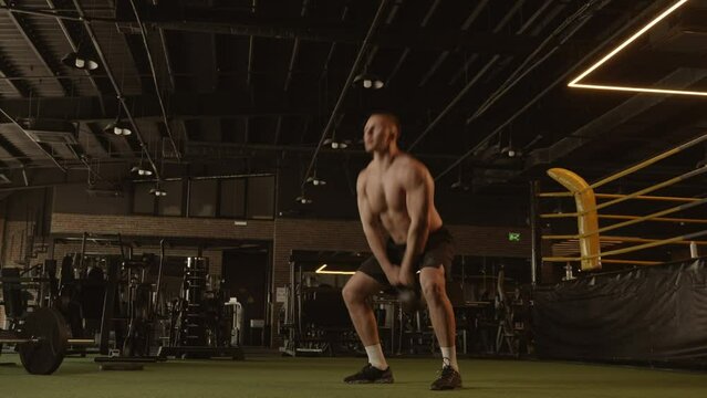 Barechested man squatting with dumbbell in a dark gym