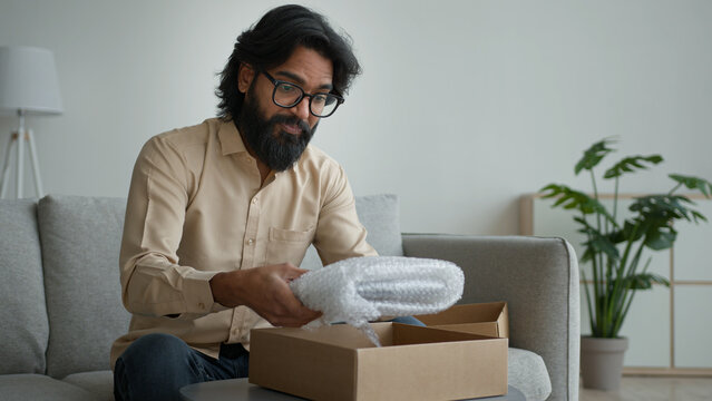 Happy male customer client consumer Arabian indian man guy in glasses open cardboard box at home unpack online shopping order purchase wrapped package get delivery present gift unpacking carton parcel