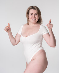 Portrait of plus size woman in bodysuit raising both hands and showing thumbs up, posing on white background. Caucasian extended sizes model is smiling and looking at camera. Body positive concept