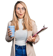 Young blonde woman wearing business style, drinking coffee and holding clipboard making fish face...