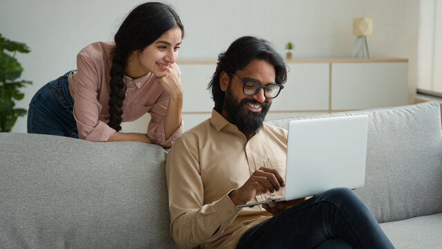 Indian muslim man husband with laptop talk to woman wife behind. Happy arabian diverse ethnic couple discuss internet order purchases choose estate apartment on website shopping online ecommerce app