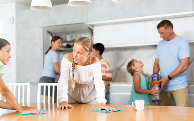 Fototapeta premium Large family is cleaning room in house - two daughters are dusting table, dad and son are vacuuming the room
