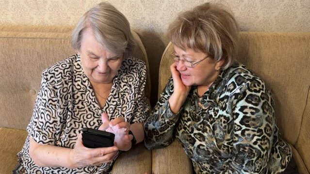 Two women sit and talk indoors. One old woman shows photos to woman using a smartphone.