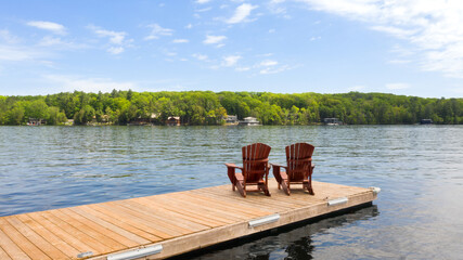 Two Adirondack chairs rest on a wooden dock in sunny Muskoka, Canada, facing a tranquil lake....