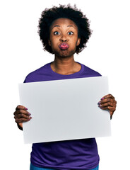 African american woman with afro hair holding blank empty banner puffing cheeks with funny face....