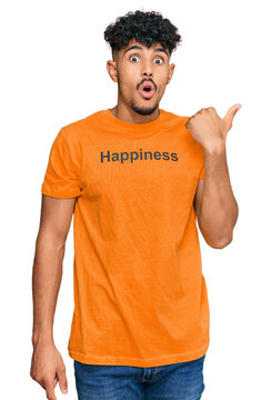 Young arab man wearing tshirt with happiness word message surprised pointing with hand finger to the side, open mouth amazed expression.