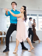 Positive emotional young guy in blue shirt enjoying impassioned merengue with female partner wearing elegant dress in latin dance class. Amateur dancing concept..