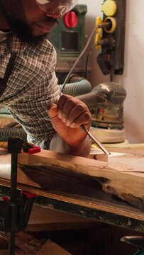 Vertical video African american sculptor shaping raw timber using chisel and hammer in carpentry shop, creating wood art, wearing safety glasses. Artist making wood sculptures, engraving lumber with