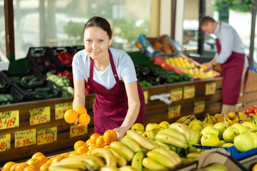 With practiced hand, woman supermarket worker arrange oranges in neat pyramid by window. Female...