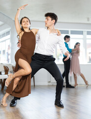 Stylish emotional young hispanic guy in white shirt and black trousers enjoying impassioned merengue with female partner wearing elegant dress in latin dance class. Amateur dancing concept