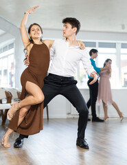 Fototapeta premium Stylish emotional young hispanic guy in white shirt and black trousers enjoying impassioned merengue with female partner wearing elegant dress in latin dance class. Amateur dancing concept