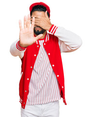 Young man with beard wearing baseball uniform covering eyes with hands and doing stop gesture with sad and fear expression. embarrassed and negative concept.