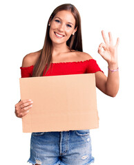 Young beautiful hispanic woman holding banner cardboard doing ok sign with fingers, smiling friendly gesturing excellent symbol